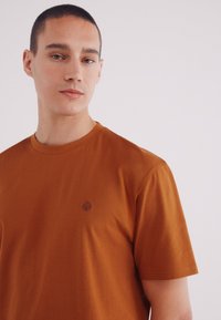 Bruine T-shirt met korte mouwen, gemaakt van zachte stof, met een klein boomlogo geborduurd op de borst. Eenvoudig en minimalistisch ontwerp.