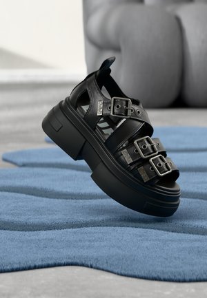 Sandale plateforme en cuir noir avec trois boucles argentées réglables, présentée sur une surface texturée bleue avec un arrière-plan flou.