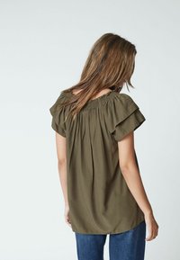 Donna con capelli lunghi indossa una blusa verde oliva a spalle scoperte con maniche a volantino e jeans blu, voltata di spalle contro uno sfondo semplice.