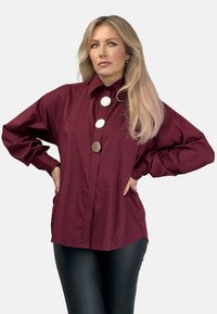 Burgundowa koszula na guziki z dużymi srebrnymi guzikami, długimi rękawami, wysokim kołnierzem i luźnym krojem z bocznymi kieszeniami. Gładka tekstura.