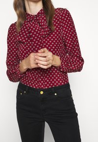 Mujer vistiendo una blusa roja con un patrón y cuello de lazo, y pantalones negros, con las manos entrelazadas a la cintura, contra un fondo blanco.