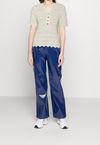 Béžový pletený top s krátkými vroubkovanými rukávy a knoflíkovým detailem, doplněný lesklými tmavě modrými širokými kalhotami a bílými sportovními botami.