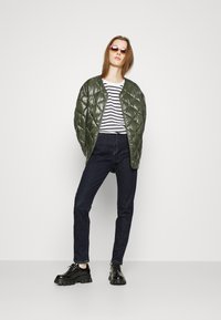 Casaco acolchoado verde-oliva, camisa riscada branca e preta, jeans escuros e sapatos pretos chunky. Modelo está em pé com as mãos na cintura.