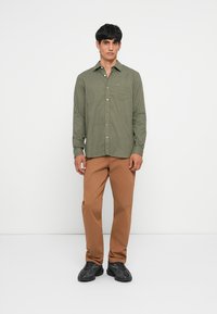Man staat tegen een witte achtergrond gekleed in een groen geruit overhemd met lange mouwen, een broek in bruin en zwarte sneakers.