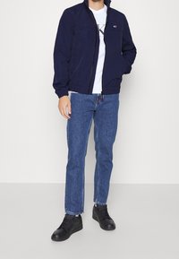 Námořnicky modrá bomber bunda s límcem na stojáka, zapínáním na zip a elastickými manžetami; v kombinaci s modrými džíny se rovnými nohavicemi a černými teniskami.