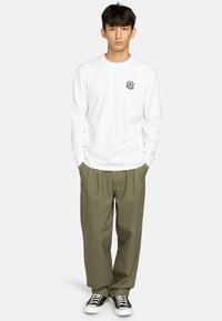 Camicia bianca a maniche lunghe con scollatura rotonda e un piccolo logo blu navy sul petto, abbinata a pantaloni larghi verde oliva e sneaker nere.