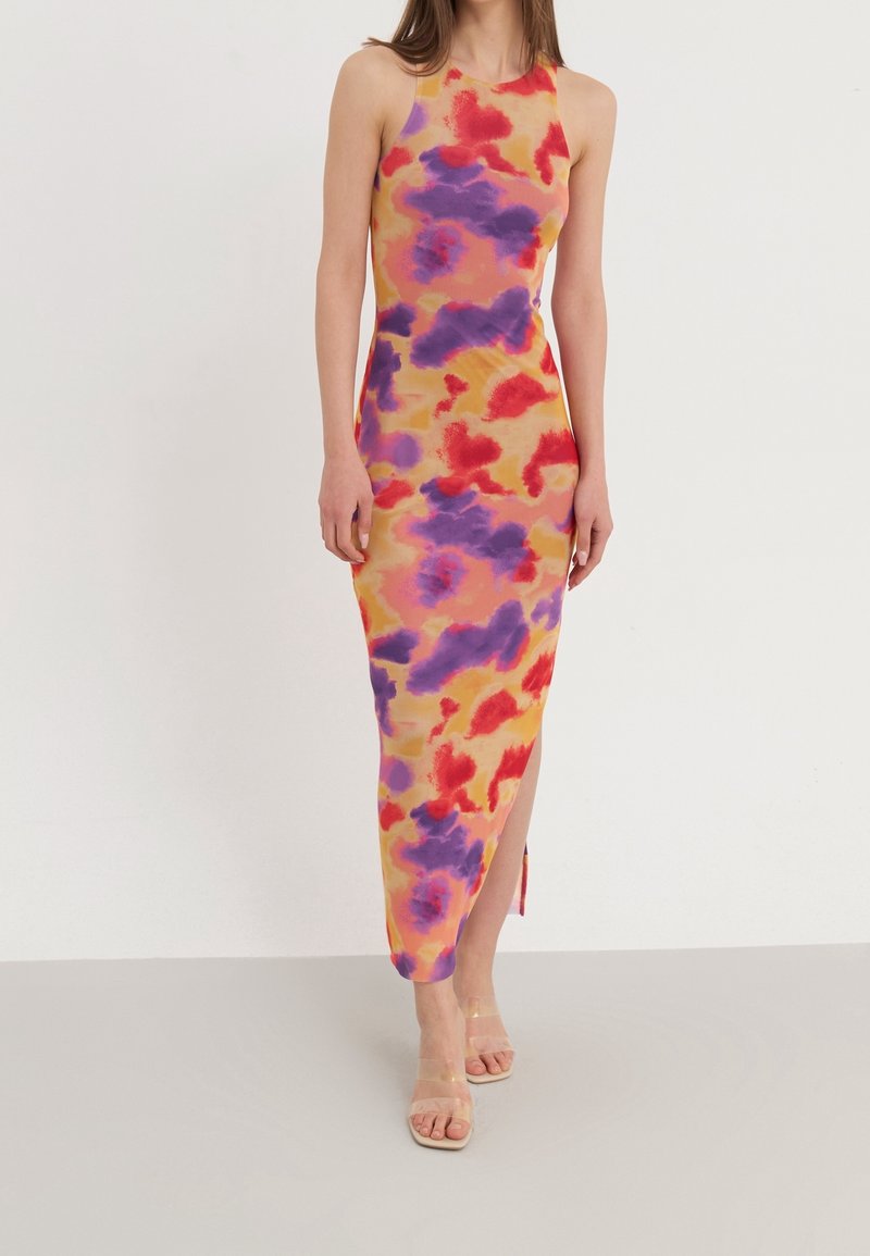 Robe longue moulante sans manches avec des motifs abstraits rouges, violets et orange, fente sur le côté, portée avec des talons ouverts à bride transparente.