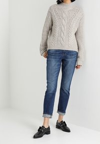 Beżowy sweter w splot warkocza z prążkowanym dołem, zestawiony z niebieskimi dżinsami z podwiniętymi nogawkami. Czarnych butów z metalowymi akcentami dopełnia stylizację.