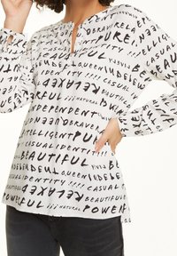 Weiße Bluse mit schwarzem handgeschriebenem Text in unterschiedlichen Größen. Das Design enthält Wörter wie "schön" und "mächtig". Lange Ärmel und ein Schlüsselloch-Nacken.