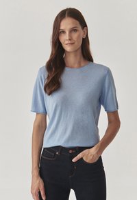 Top tricotat cu mânecă scurtă de culoare albastru deschis, cu o croială lejeră și guler rotund. Este asortat cu o pereche de blugi negri din denim, cu cusături contrastante și buzunare.