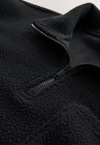 Čierna fleecová bunda s čiastočne zapnutým predným zipsom a stojacím golierom, zobrazujúca textúrovanú, mäkkú látku zblízka.