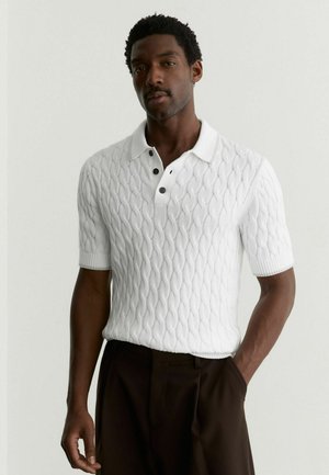 Muž v bílé krátkosleevé kabelové pletené polokošili a tmavých kalhotách, stojící s jednou rukou v kapse na jednobarevném pozadí.