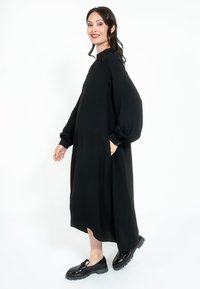 Crna midi haljina s dugim, obimnim rukavima i bočnim džepovima, napravljena od glatkog materijala. Uparena s sjajnim crnim cipelama bez pertli.