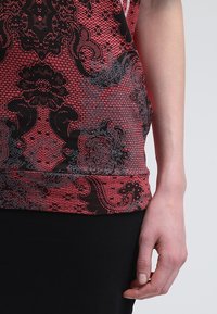 Haut rouge sans manches avec des motifs floraux complexes noirs et gris, rappelant de la dentelle, porté sur le torse et le bras droit d'une personne, sur un fond uni.