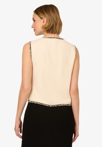 Hedvábný krémový top bez rukávů s texturovanou tkaninou, černým lemem na výstřihu a jemnými černými akcenty na spodním lemu, nošený přes černou sukni.