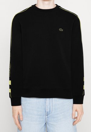 Sweat-shirt noir à manches longues avec des rayures jaunes sur les manches et un petit logo crocodile sur la poitrine gauche, porté avec un jean bleu clair.