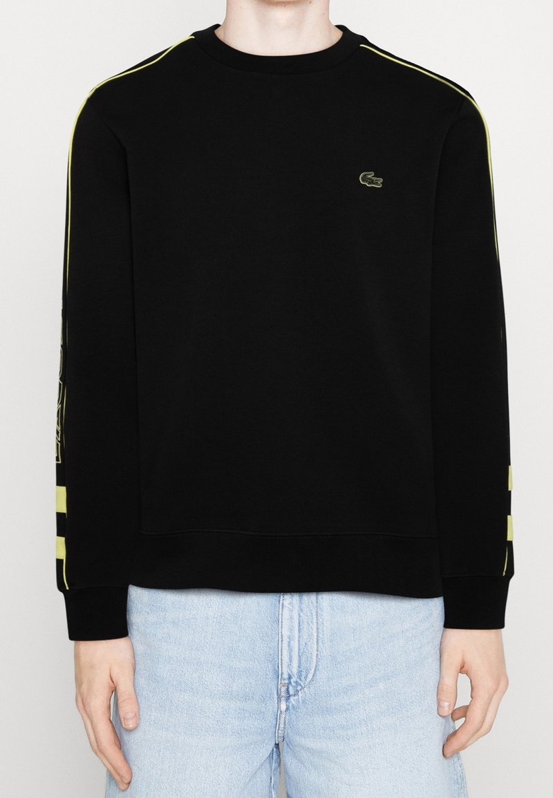Sweat-shirt noir à manches longues avec des rayures jaunes sur les manches et un petit logo crocodile sur la poitrine gauche, porté avec un jean bleu clair.