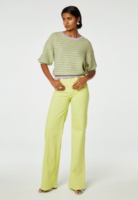 Gebreide, kortef op mouw trui met een groen en paars patroon, gecombineerd met lichtgroene, hoge taille, wijde broek. Schoenen met zilveren accenten.