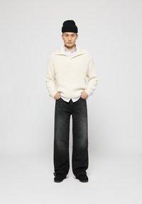 Jersey de punto color crema con un cuello de cremallera de un cuarto, superpuesto a una camisa a rayas. Jeans de pierna ancha en gris oscuro y zapatos negros de slip-on.