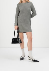 Šedé pletené svetrdresses s dlouhými rukávy, doplněné černou kabelkou a špičatými černými balerínami. Model má na sobě světle šedé žebrované ponožky.