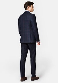 Granatowa marynarka w kratkę z czerwonymi liniami, dopasowany krój, pojedynczy rozporek, noszona z pasującymi spodniami i brązowymi butami.