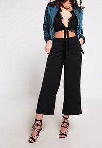 Žena v černém krajkovém topu, tyrkysové bomberové bundě, černých širokých kalhotách a černých krajkových sandálech na vysokém podpatku, stojící na bílém pozadí.