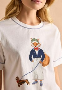 Valge puuvillane T-särk värvilise rebase graafikaga, kes kannab sinist jakki ja rohelist korki ning hoiab rihma, mis on kinnitatud pruuni taksikoera külge.