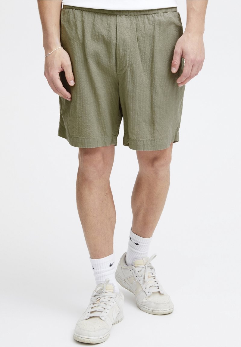 Homme portant un short texturé olive, des chaussettes blanches Nike et des baskets blanches Nike, se tenant devant un fond blanc.