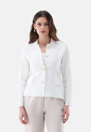 Donna che indossa un blazer bianco abbottonato sopra una maglia bianca e pantaloni beige, in piedi davanti a uno sfondo grigio chiaro uniforme.