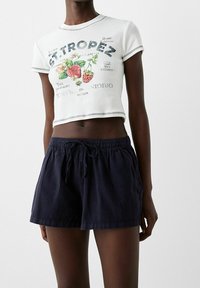 Bílý zkrácený tričko s ovocnou grafikou a textem; spárováno s tmavě námořnickými šortkami s elastickým pasem a šňůrkou na stažení.