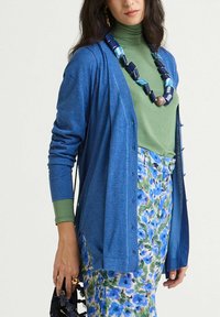 Cardigan bleu en tissu doux avec des boutons sur le devant, porté sur un haut vert, associé à une jupe à motif floral et un collier épais.