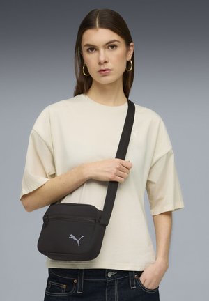 Bolso bandolera de tela negra con cierre de cremallera, con el logo de Puma. Llevado con una camiseta oversize de color beige claro y vaqueros oscuros.