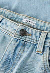 Svijetloplave traperice s izblijedjelim detaljima, vidljivim šavovima i crnim metalnim gumbom s gravurom "TWO SOON" na pojasu.