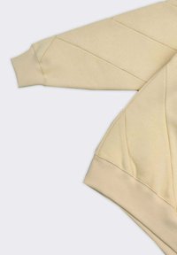 Manica di felpa beige con polsino a coste e dettagli a cucitura diagonale su tessuto liscio, posizionata su uno sfondo grigio chiaro.