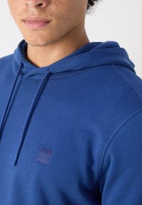 Jeune personne avec des taches de rousseur portant un sweat à capuche bleu avec des cordons de serrage et un petit logo "BOSS" sur la poitrine.