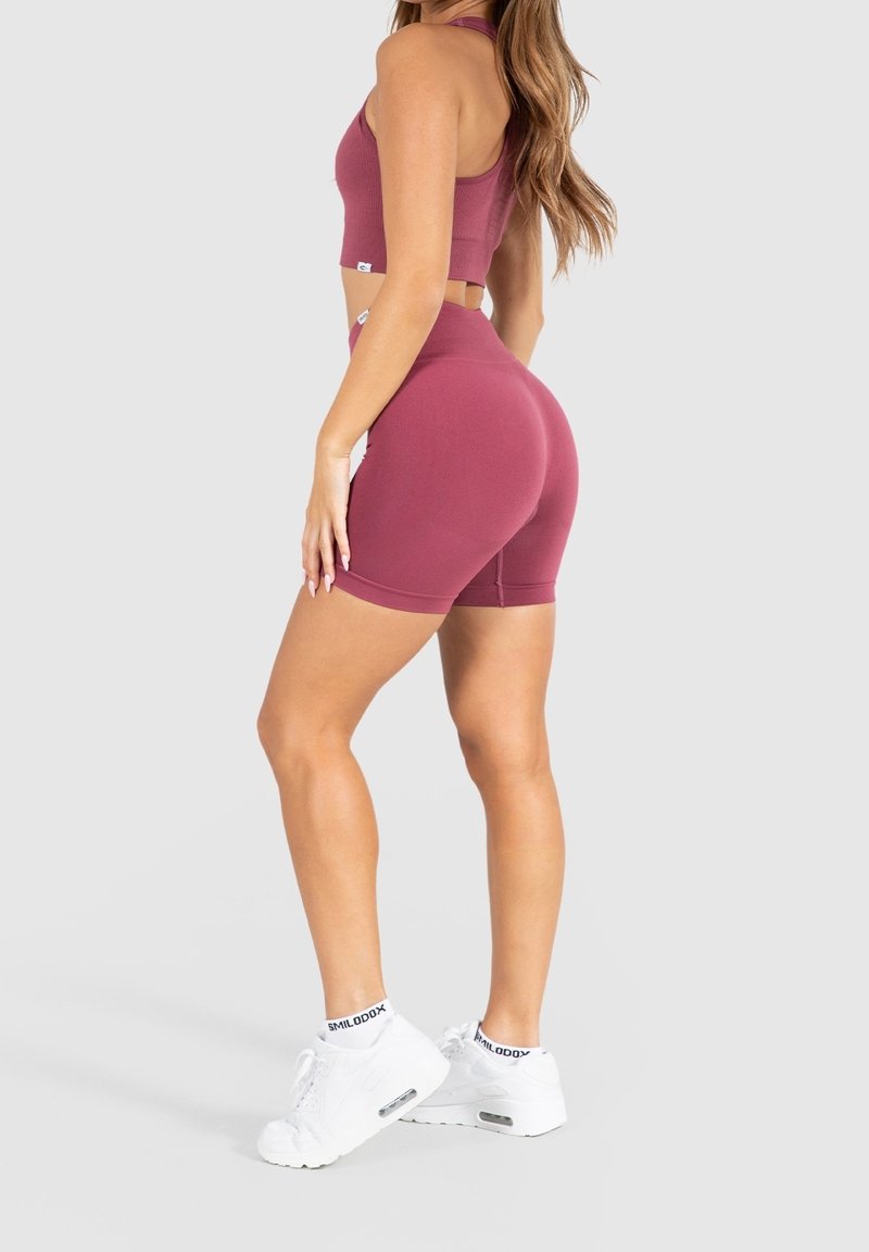 Top corto sportivo bordeaux e pantaloni shorts, tessuto a costine, design aderente, abbinato a sneakers bianche con accenti neri, visto da dietro.