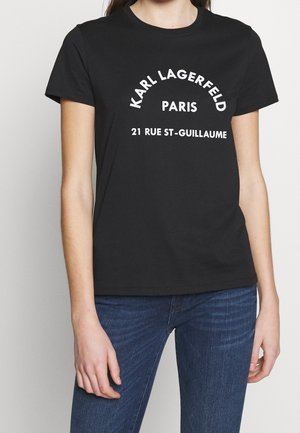 Czarny t-shirt z białym napisem "Karl Lagerfeld Paris 21 Rue St-Guillaume", noszony przez osobę w niebieskich dżinsach na jednolitym tle.