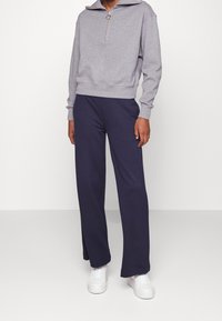Felpa grigia con zip, colletto alto e orlo a costine, abbinata a pantaloni ampi blu navy. Indossata con sneakers bianche.