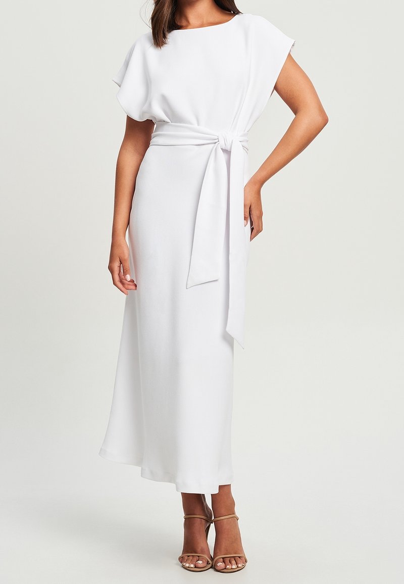 Femme portant une robe midi blanche à manches courtes avec une ceinture à la taille et des sandales beiges à talons ouverts, sur un fond uni.