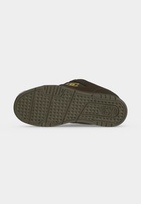 Semelle d'une basket vert olive foncé avec un motif de bande de roulement texturé et un logo près du talon sur un fond gris clair uni.