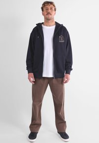 Hoodie zippé bleu marine avec poches avant et logo, associé à un pantalon beige et des baskets noires, porté sur un t-shirt blanc.