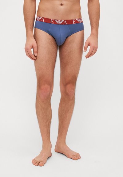 Slips pour hommes en bleu et rouge. Matériau en coton, design ajusté, avec une large ceinture affichant un logo. Texture lisse, coupe allant jusqu'à l'avant.