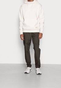 Cremefarbener Hoodie mit Fronttasche und Kordelzug, kombiniert mit dunkelgrünen Cargo-Hosen mit Seitentaschen. Schuhe: weiße und graue Sneakers.