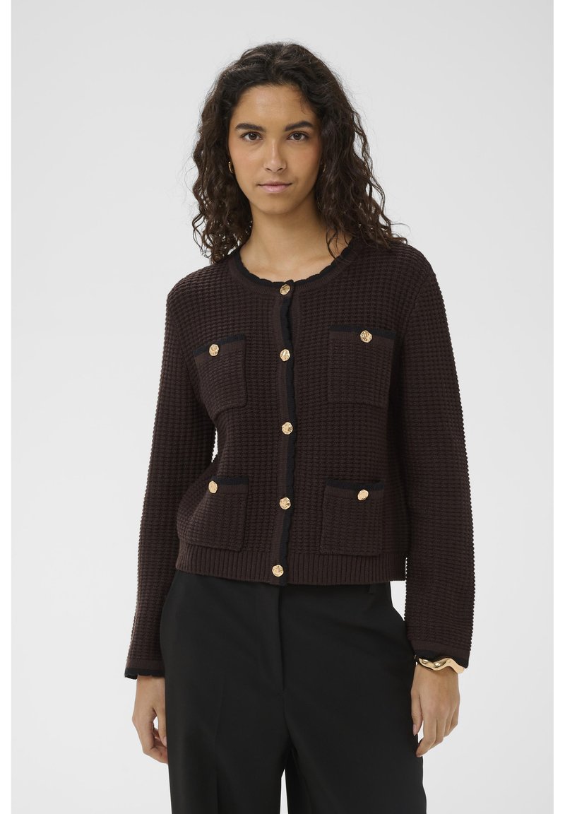 Femme aux cheveux bouclés foncés porte un cardigan marron foncé texturé avec des boutons dorés et un bord noir, associé à un pantalon noir.