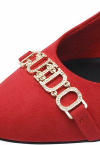 Röd mockaskinnssko med spetsig tå, med en guld kedjedetalj och glittrande detaljer. Slät textur med svart inredning.