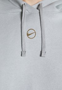 Ljusgrå hoodie med texturerat tyg, dragsnören och en metallisk guldig cirkulär logotyp med en swoosh-design framtill.