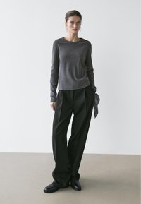 Femme aux cheveux foncés portant un haut gris à manches longues et un pantalon noir ample, tenant un tissu sombre à motifs, debout devant un fond uni.