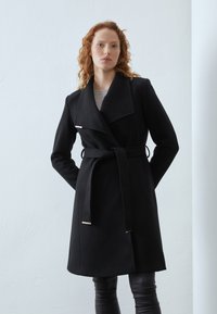 Cappotto nero con rever ampio, vita segnato da una cintura e dettagli metallici. Presenta maniche lunghe e una linea diritta, realizzato in un tessuto morbido.
