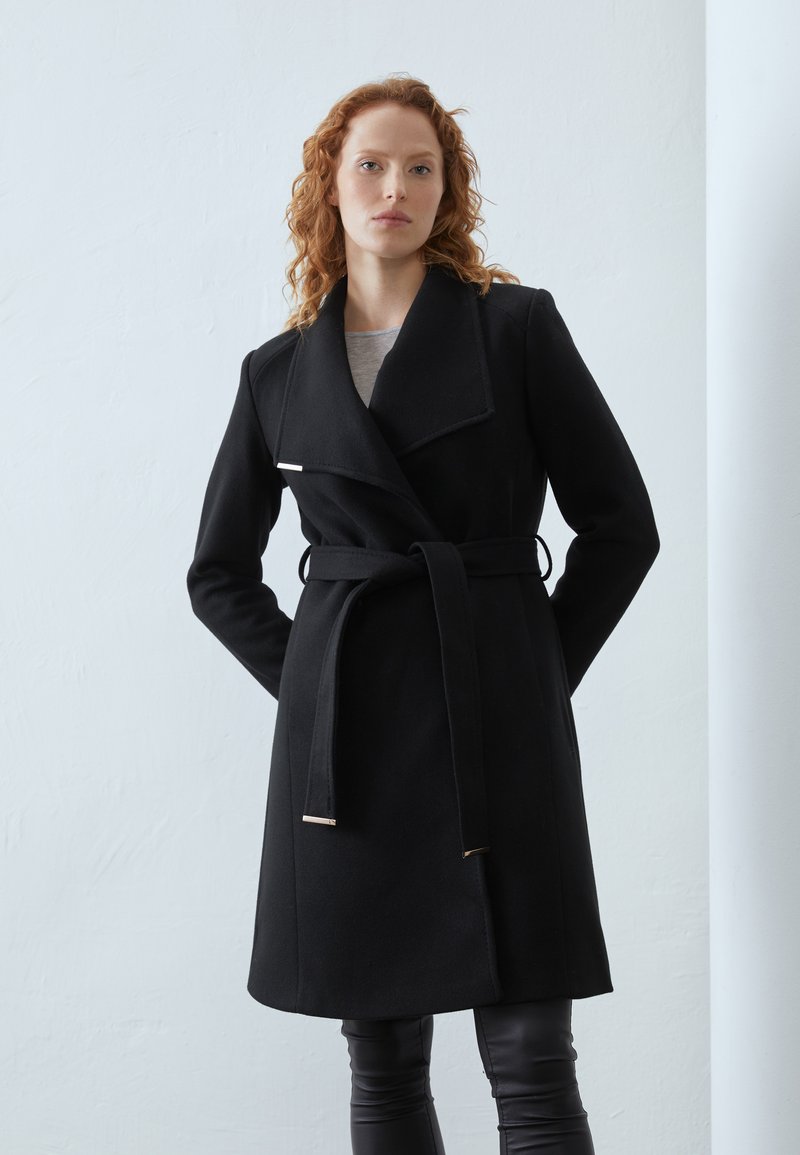 Cappotto nero con rever ampio, vita segnato da una cintura e dettagli metallici. Presenta maniche lunghe e una linea diritta, realizzato in un tessuto morbido.