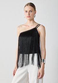 Svart frynsetopp med asymmetrisk design, enkelt stroppe og flere frynser. Kombinert med hvite vide bukser og et silver armbånd.